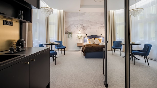 A modern hotel room with a bed, a desk with a coffee maker, and a view of a cityscape through large windows. - Astoria Boutique Suites - by BQA (Budapest)