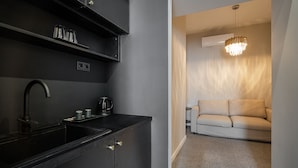 A modern kitchen with black cabinetry, a sink, and a kettle. There is a beige sofa in the adjacent living area with a chandelier above. - Astoria Boutique Suites - by BQA (Budapest)