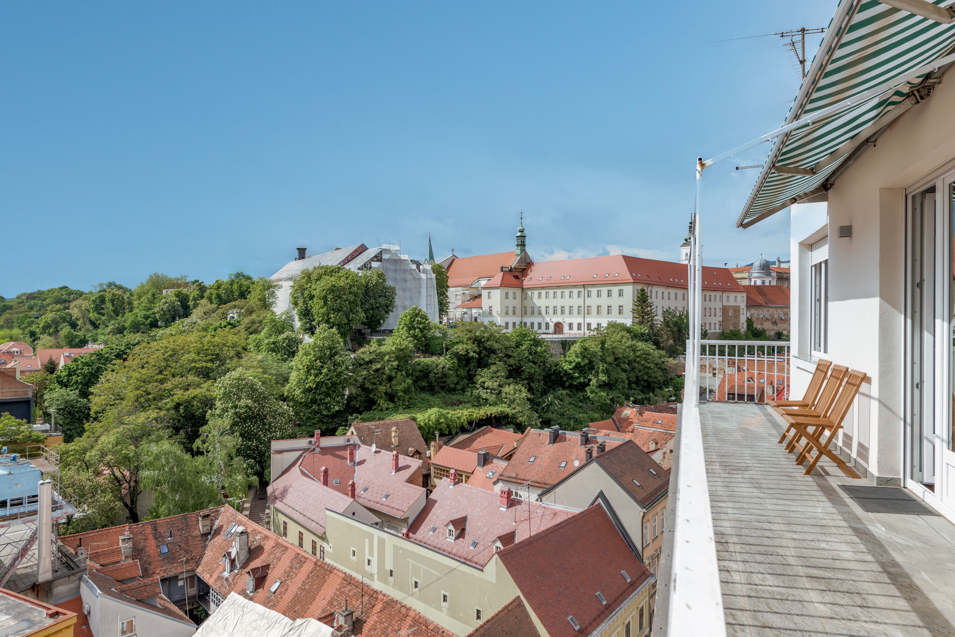 A balcony with wooden chairs, a view of red-roofed buildings, and a large building with a green spire in the background.