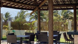 An outdoor patio area with a tiled roof, wicker furniture, and a pool in the background. - Summer Ville Itacimirim (Camaçari)