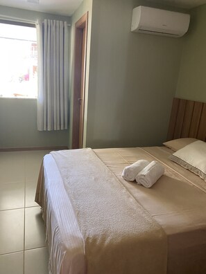 Ett hotellrum med en säng, ett fönster med gardiner, en luftkonditioneringsanläggning och ett klinkergolv.