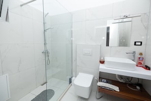 A modern bathroom with a glass-enclosed shower, a white toilet, a sink with a mirror, and a shelf with toiletries.