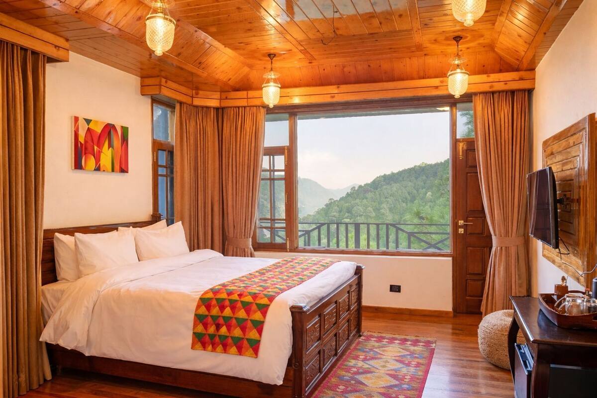A bedroom with a large bed, wooden ceiling, a window with a view of greenery, and a mounted TV.