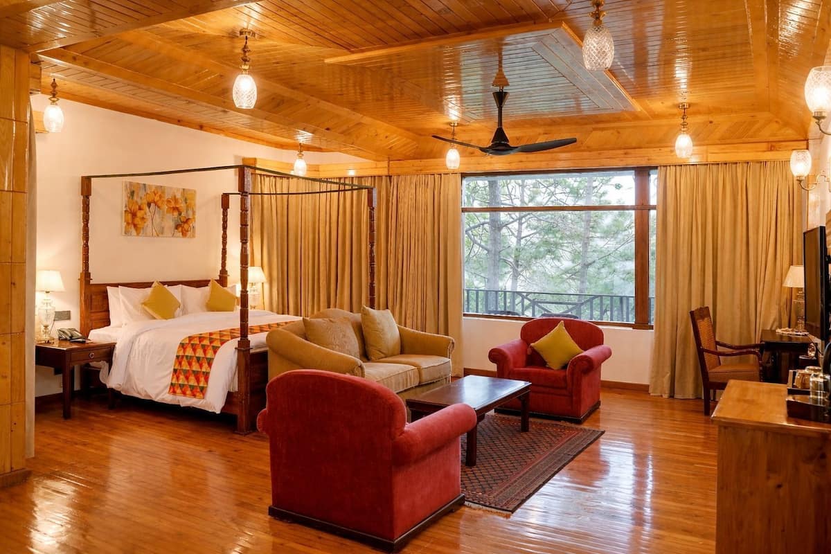 A four-poster bed, a sofa, a red armchair, a wooden coffee table, and a television in a room with wooden floors and a large window.