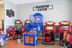 Zona de juegos con diversas máquinas de arcade, incluyendo baloncesto y juegos de carreras, y un letrero de neón en la pared.