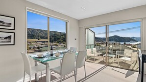 Un espace repas moderne avec une table en verre et des chaises blanches, offrant une vue panoramique sur des collines et un plan d’eau.