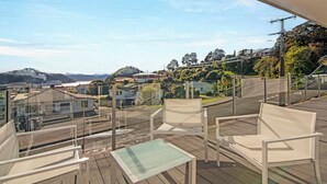 Un balcon avec des chaises blanches et une table en verre, donnant sur un quartier résidentiel avec des maisons et une colline au loin.