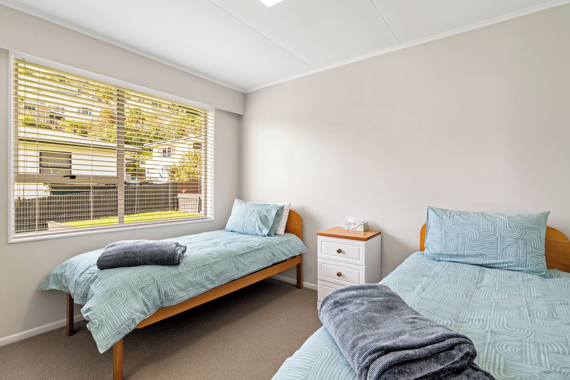 A bedroom with two beds, a window with blinds, a bedside table, and a carpeted floor.