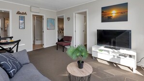 Un salon avec un canapé, un téléviseur posé sur un meuble blanc, une plante en pot et un tableau représentant un coucher de soleil.