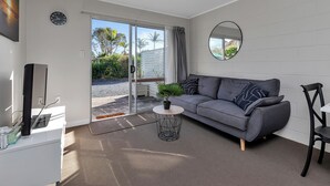 Un salon moderne avec un canapé gris, une table basse ronde ornée d’une plante, un téléviseur posé sur un meuble blanc et une porte-fenêtre coulissante donnant sur un balcon.