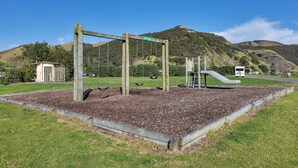 Une aire de jeux avec des balançoires, un toboggan et une structure d’escalade, entourée d’une pelouse et d’une clôture en bois.