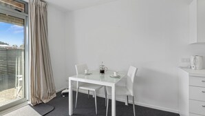 A modern hotel room with a glass table, white chairs, a coffee maker, and a view of the city through a large window.