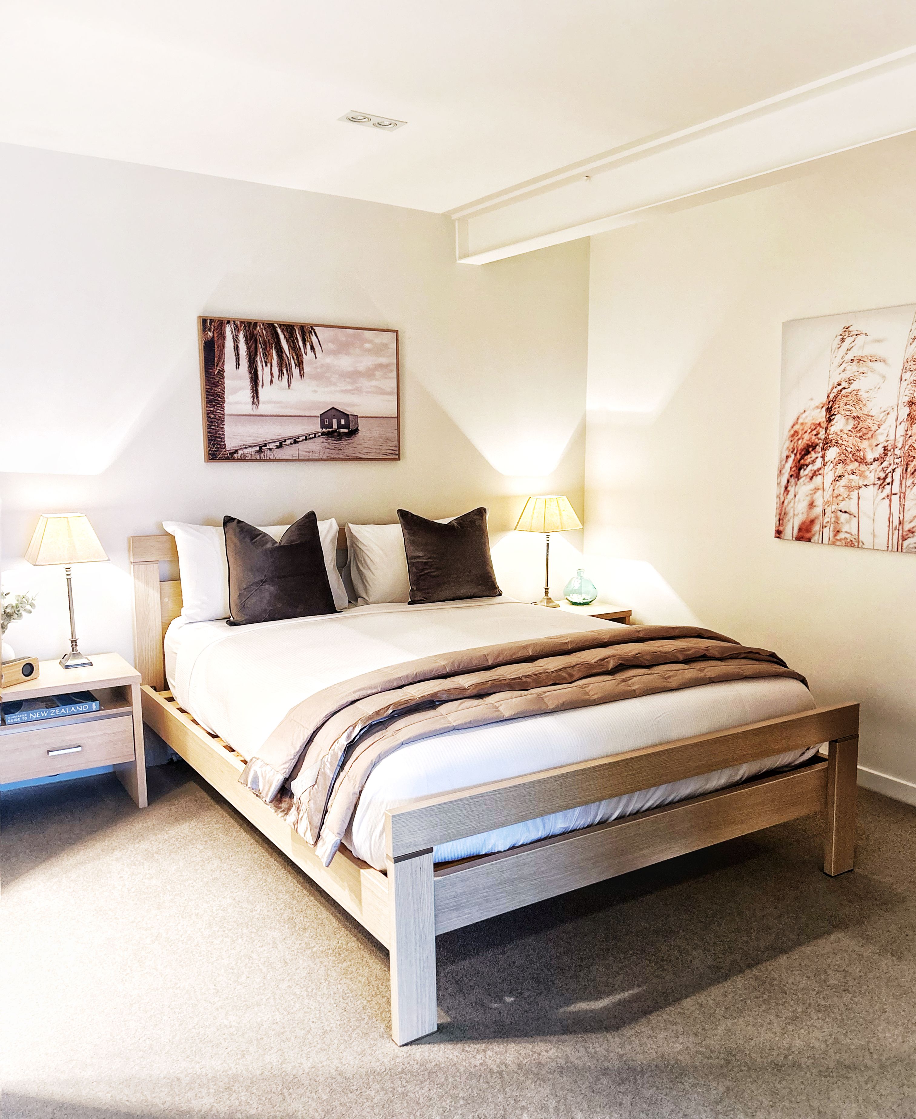 A bedroom with a wooden bed, two bedside tables, a nightstand, a lamp, a framed picture on the wall, and a carpeted floor.