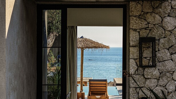 Eine Steinsäule führt zu einer Terrasse mit einem Holzstuhl und einem Reedschirm, mit Blick auf ein Schwimmbecken und das Meer.