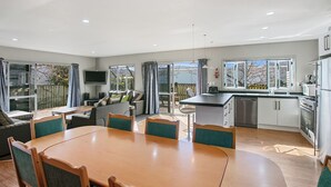 A modern kitchen with a dining area, large windows, and a flat-screen TV. - Family Harmony - Waihi Beach Holiday Home (Waihi Beach)