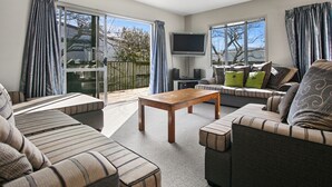 A spacious living room with a large flat-screen TV, a wooden coffee table, and a sofa with striped cushions. There are sliding glass doors leading to a balcony. - Family Harmony - Waihi Beach Holiday Home (Waihi Beach)