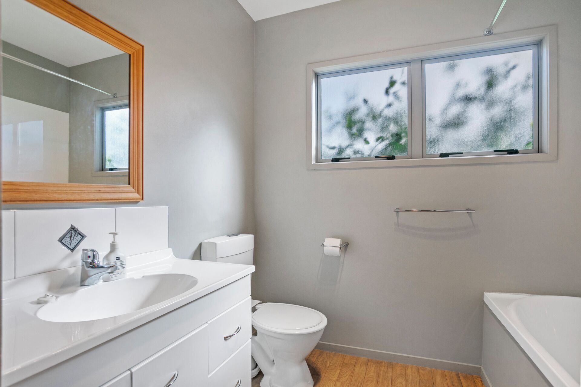 Un baño moderno con lavamanos, inodoro y bañera blancos. Hay un ventanal sobre el inodoro y un espejo sobre el lavamanos.