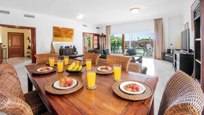 Un comedor con una mesa de madera preparada para el desayuno, sillas de mimbre y vistas al exterior.