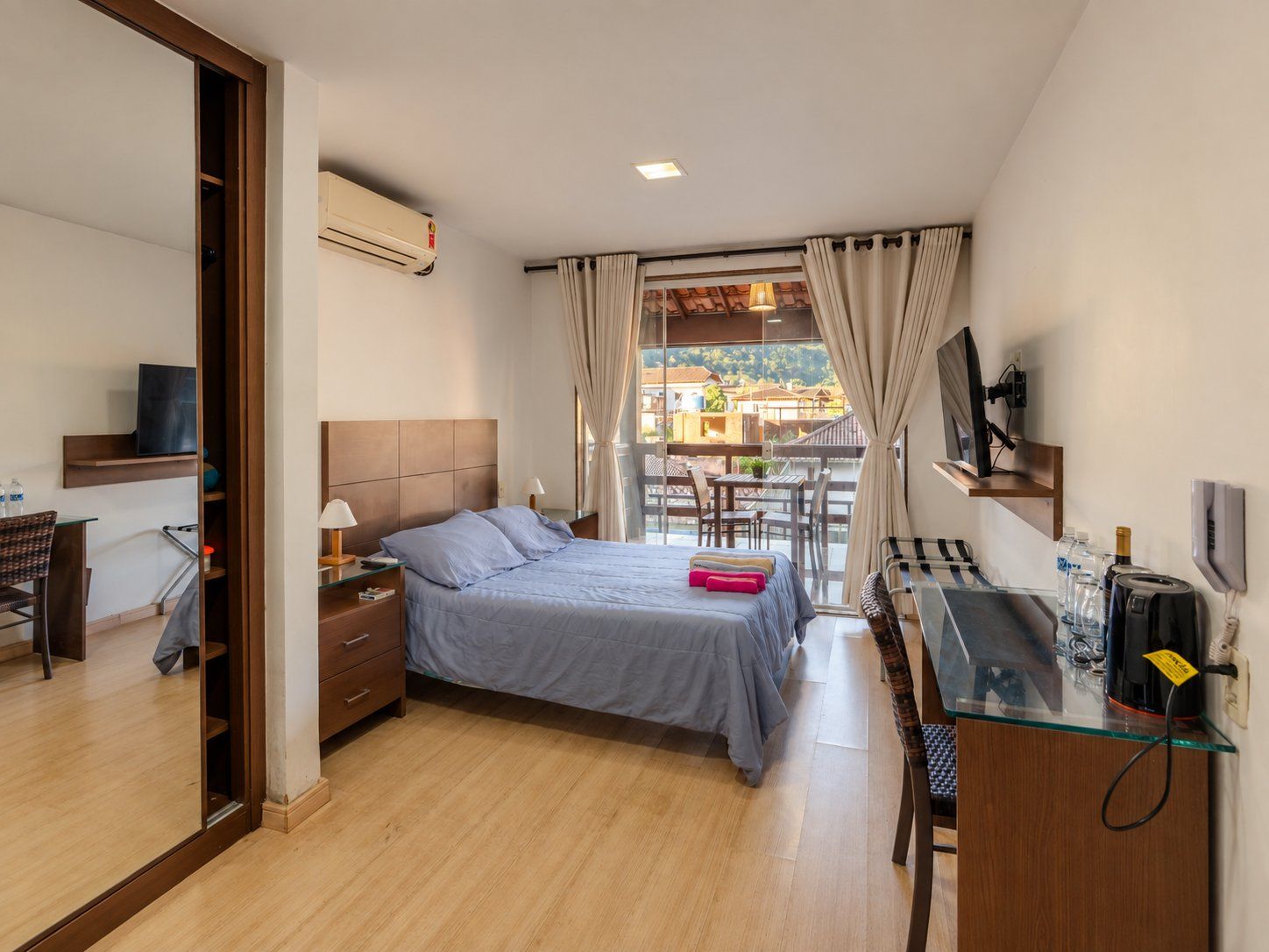 Hotelkamer met een bed, nachtkastje, lamp en bureau met stoel. Er is een raam met gordijnen, uitzicht op gebouwen buiten en een airconditioningunit aan de muur.