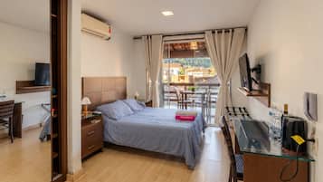 Hotelkamer met een bed, nachtkastje, lamp en bureau met stoel. Er is een raam met gordijnen, uitzicht op gebouwen buiten en een airconditioningunit aan de muur.