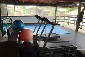 Uma academia com equipamentos de ginástica, incluindo esteira e bolas de exercício, e vista para edifícios externos.