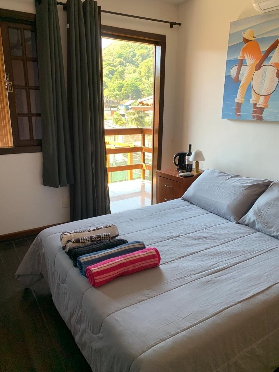 Un lit bien fait, avec une serviette à rayures, une table de chevet en bois sur laquelle se trouve une lampe, et un tableau représentant deux personnes sur un balcon.