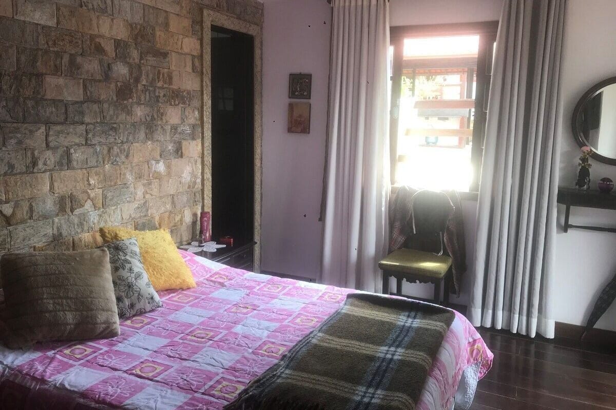 Un dormitorio con cama, una silla, ventana con cortinas y pared de piedra.