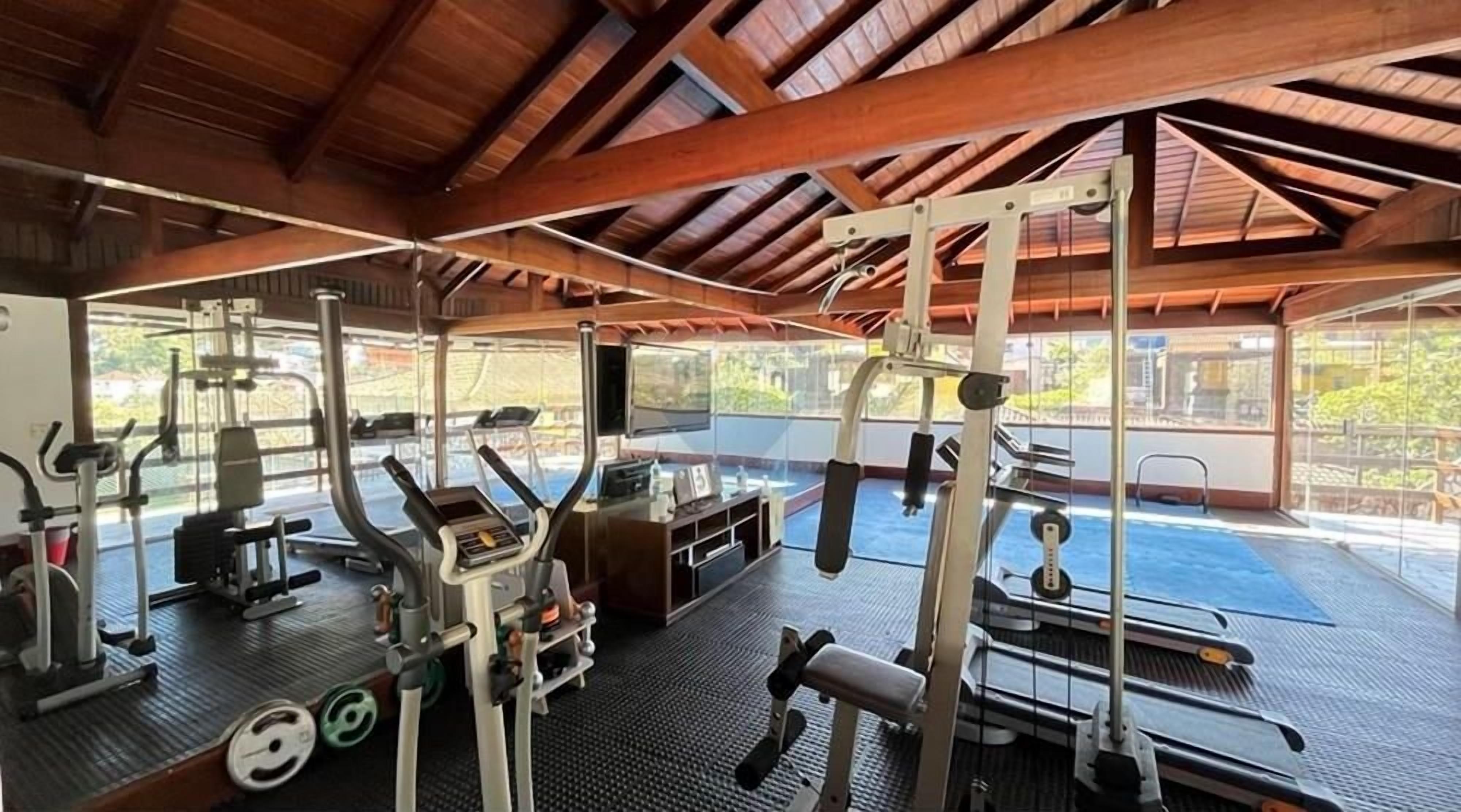 Uma academia com diversos equipamentos de exercícios, incluindo esteiras e aparelhos de musculação, sob um telhado de madeira com grandes janelas.