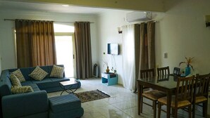 A living room with a sofa, ottoman, and a dining table with chairs. There is a television on the wall and an air conditioning unit. - WHITE VILLA No. 8
 (Hurghada)