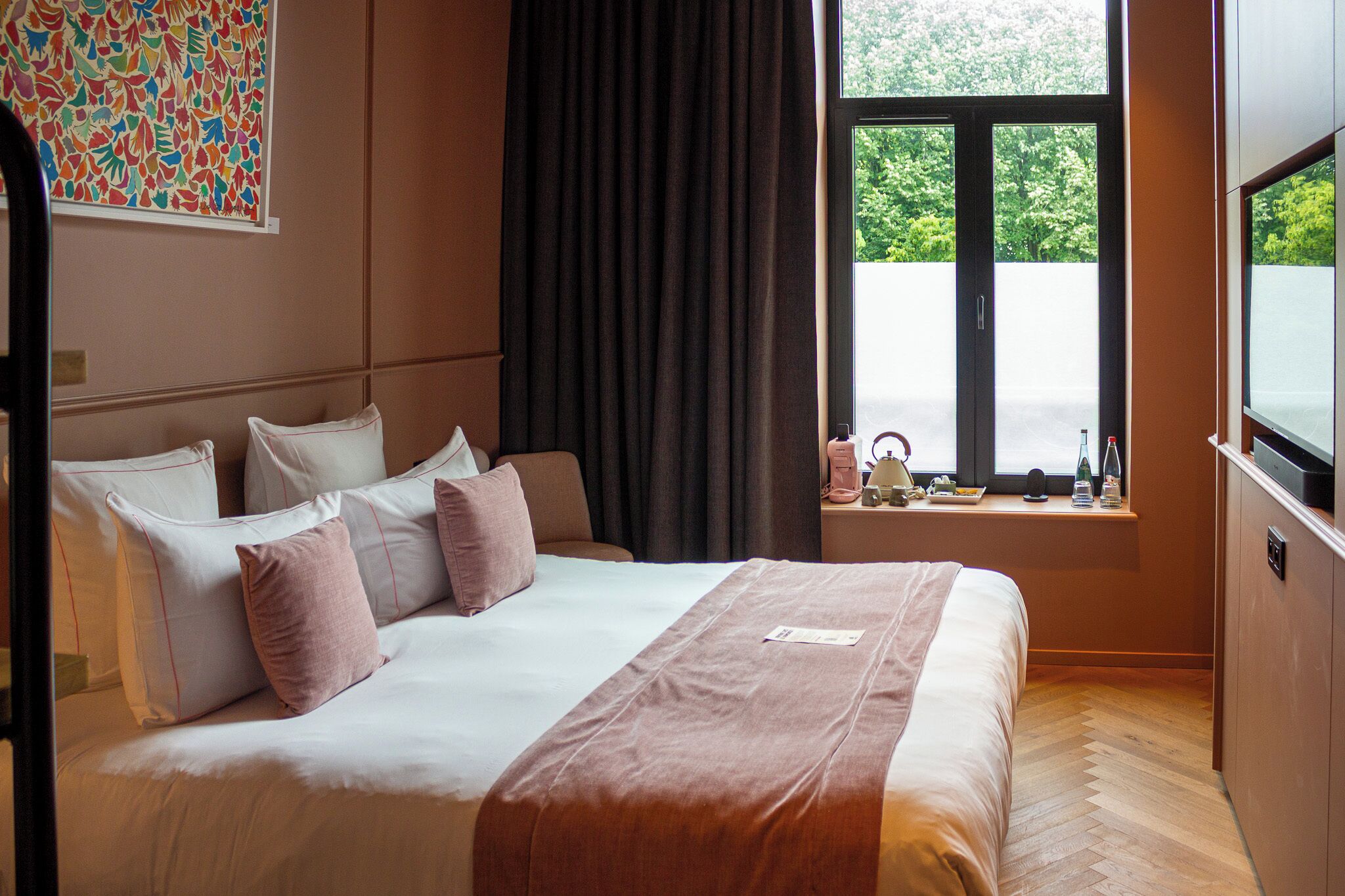 A hotel room with a large bed, a window with a view of greenery, and a television on a stand.
