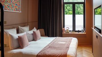 A hotel room with a large bed, a window with a view of greenery, and a television on a stand.