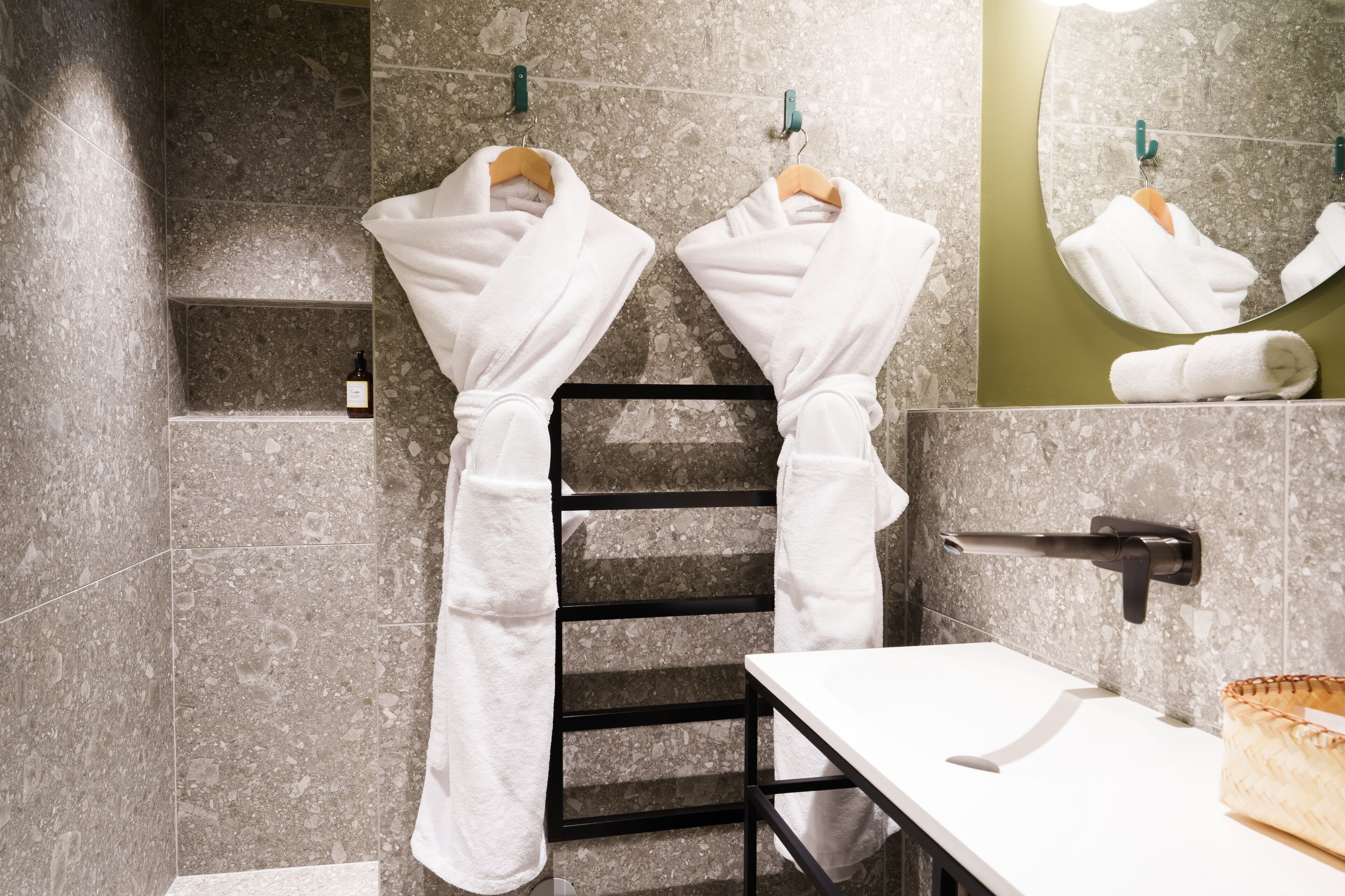 Un baño con dos batas blancas colgadas en un perchero, un espejo, un perchero para toallas y un lavabo con una toalla sobre el mostrador.