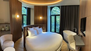 Una habitación de hotel moderna con una cama grande, un escritorio y una silla. Hay dos ventanas con cortinas y un televisor montado en la pared.
