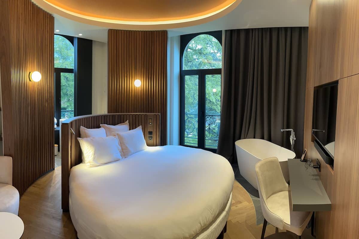 Una habitación de hotel moderna con una cama grande, un escritorio y una silla. Hay dos ventanas con cortinas y un televisor montado en la pared.