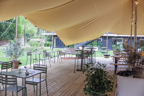Un patio exterior con mesas y sillas, un toldo grande y vistas del jardín circundante.