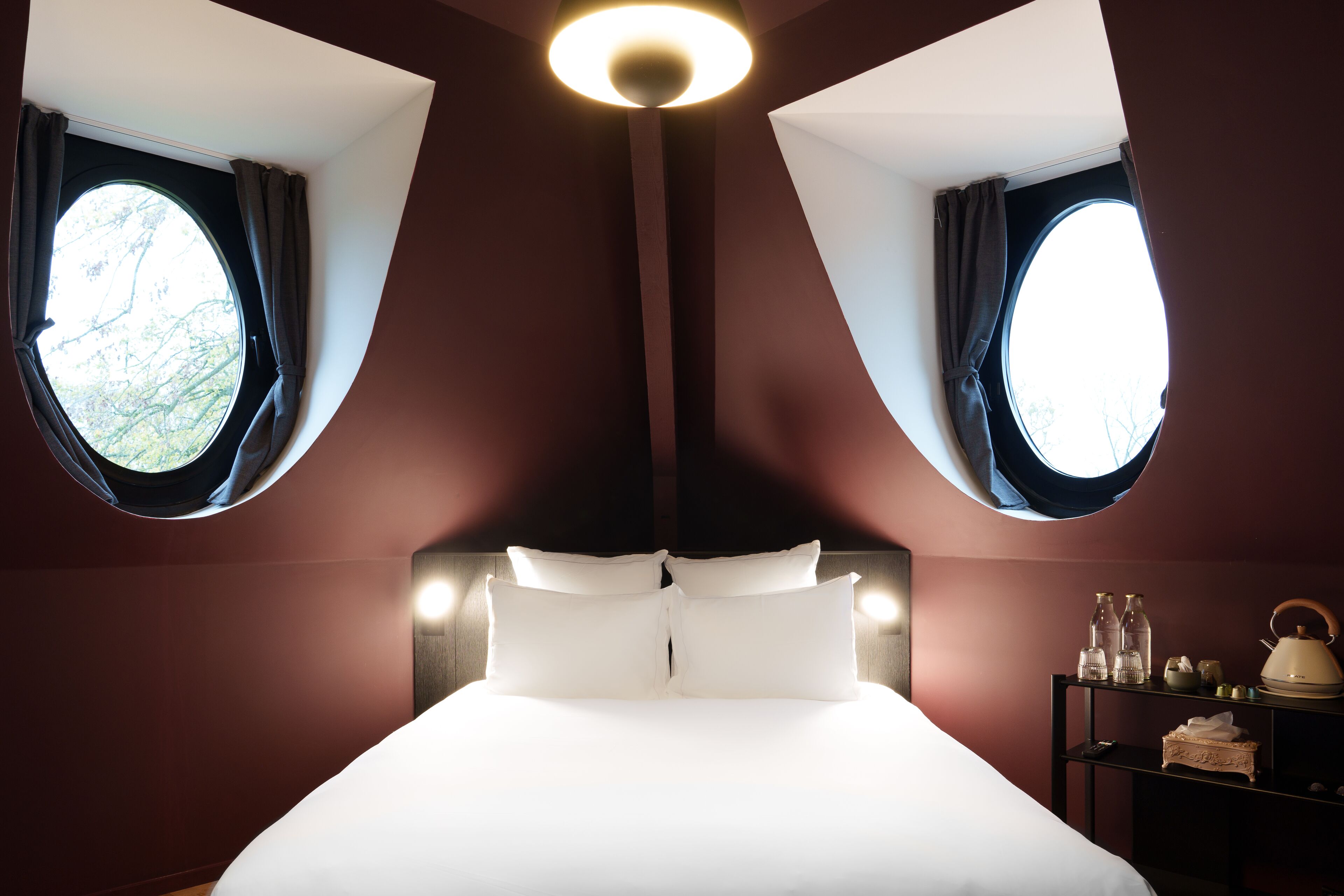 Una habitación con una cama grande, dos ventanas con cortinas, una mesita de noche con hervidor y una repisa con botellas y una cesta.