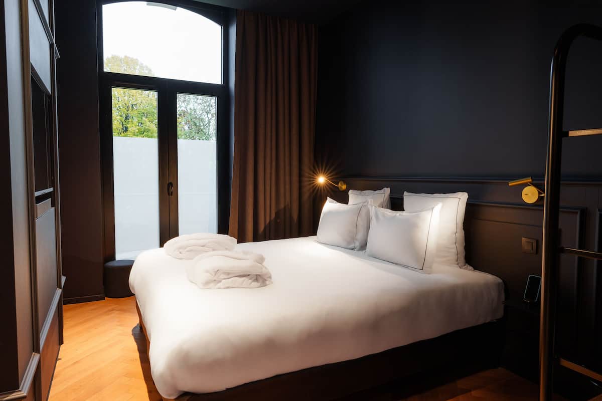 Una cama bien hecha con sábanas y almohadas blancas, una mesita de noche con lámpara, y vistas al verdor a través de un ventanal grande.