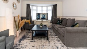 Uma sala de estar moderna com uma TV de tela plana, dois sofás, uma mesa de centro e espelhos decorativos nas paredes.