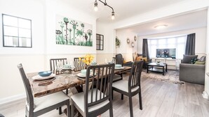 Uma sala de estar moderna com área de jantar, apresentando uma mesa de jantar de madeira, cadeiras escuras, um sofá cinza e uma TV.