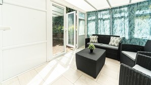 Pátio iluminado pelo sol com porta de vidro deslizante para um jardim, um conjunto de sofá de vime preto e uma pequena mesa com uma planta.