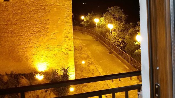 Varanda com vista para uma rua à noite, com um edifício de pedra de um lado e um carro estacionado abaixo.