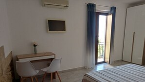 Quarto com cama, escrivaninha com duas cadeiras, televisão pequena e janela com cortinas.