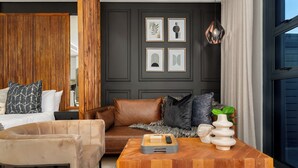 Een moderne hotelkamer met een donkere muur, een houten roomdivider, een bruine leren bank, een beige fauteuil, een houten bijzettafeltje met een afstandsbediening, en kunstwerken in lijsten aan de muur.