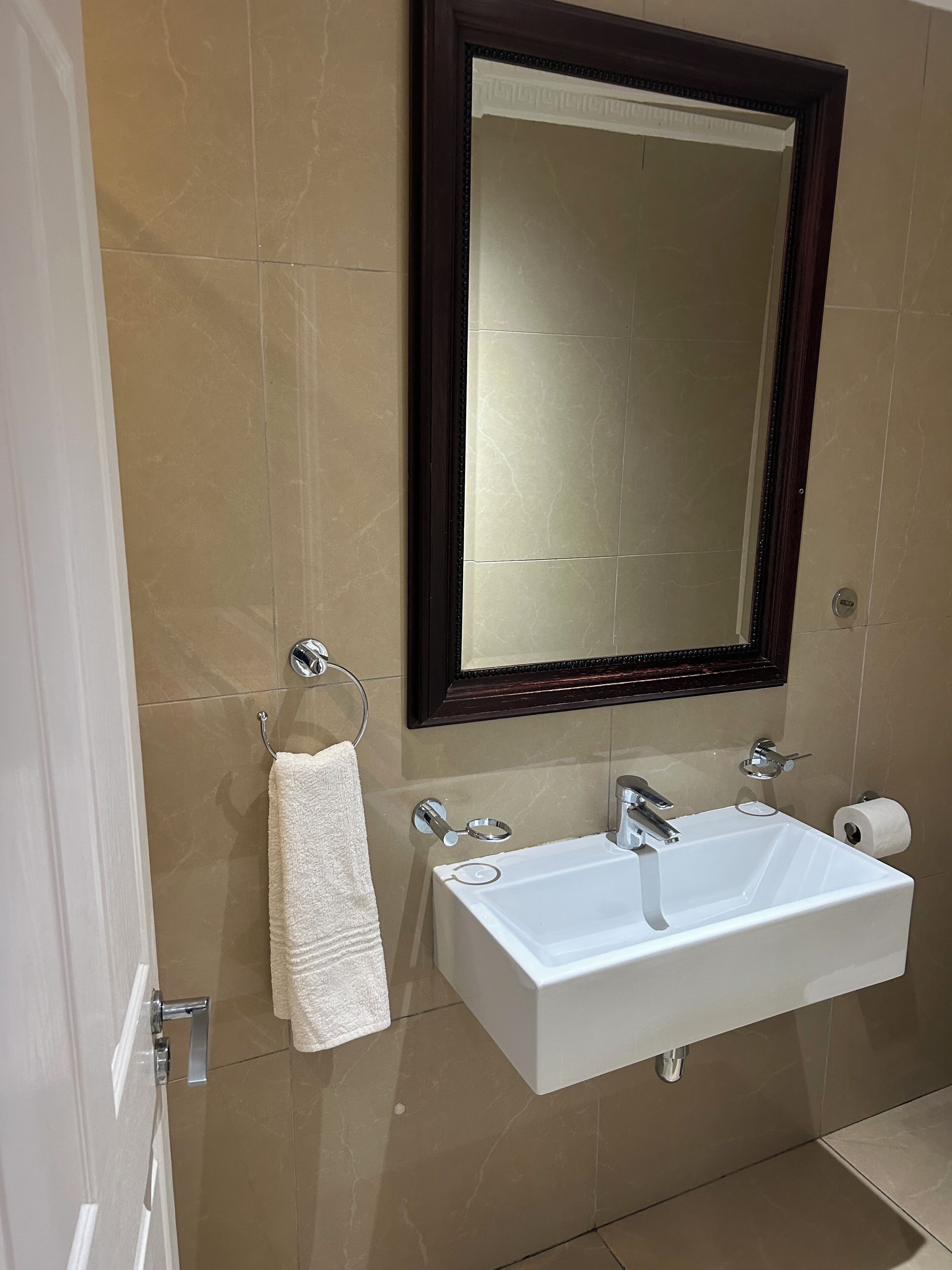 Et badeværelse med en hvid vask, et håndklædetørrestativ med et hvidt håndklæde, et spejl med mørk ramme og en toiletpapirholder.