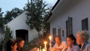 Een groep mensen dineert buiten aan een tafel met kaarsen, omgeven door een wit gebouw en bomen.