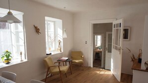 Een lichte kamer met houten vloeren, een eettafel, twee stoelen, een klein tafeltje met boeken en een deur naar een andere kamer.
