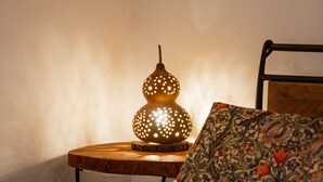 A neatly made bed with a rolled white towel, a decorative lamp, and a floral pillow on a wooden nightstand.