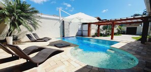 Una piscina con sillas de descanso y una pérgola, rodeada por un jardín ajardinado y un edificio con fachada de piedra.