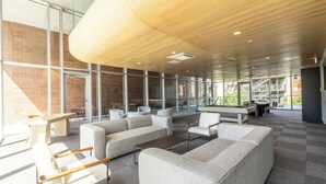A modern lounge area with sofas, chairs, and tables, large windows, and a curved wooden ceiling.