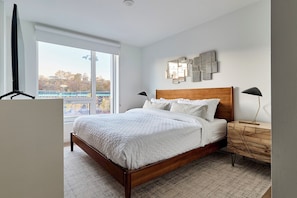 Quarto com cama de madeira, roupa de cama branca, mesa de cabeceira, abajur e vista para o exterior através de uma janela grande.